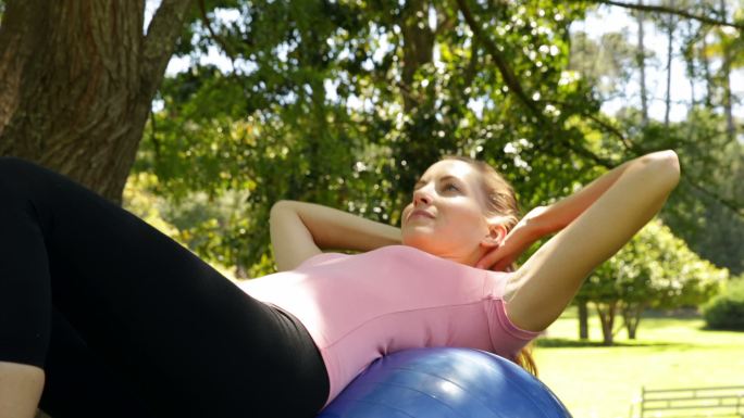 在一个阳光明媚的日子里，健身的女人在公园里的健身球上做仰卧起坐