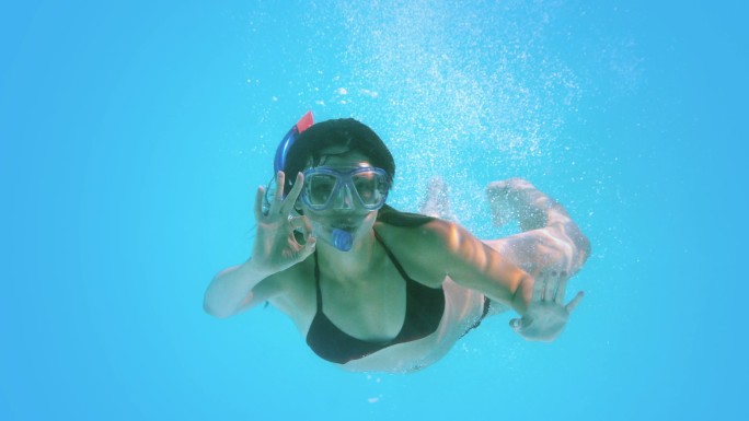 黑发女子在水下游泳，戴着通气管，慢动作做ok手势
