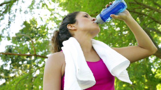 在一个阳光明媚的日子里，一名妇女在公园里锻炼后，擦拭她的额头和喝水