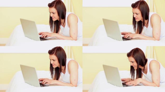 漂亮的黑发女子躺在床上用她的笔记本电脑在卧室在家里