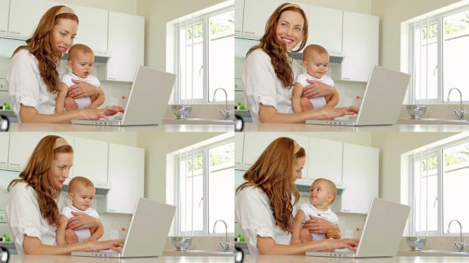 漂亮的黑发女子使用笔记本电脑，抱着她的宝贝女儿在厨房里