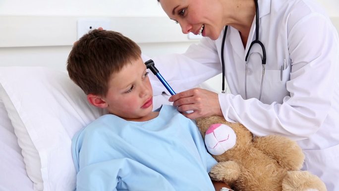 在医院病房的病床上，医生正在检查一个生病的小男孩的耳朵