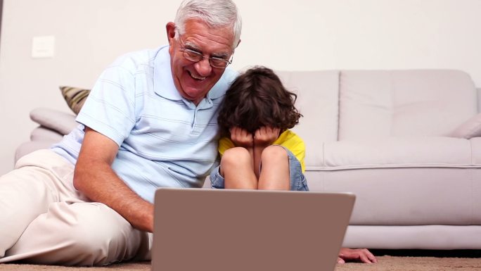 一位老人和他的孙子坐在地板上，在家里的客厅里使用笔记本电脑