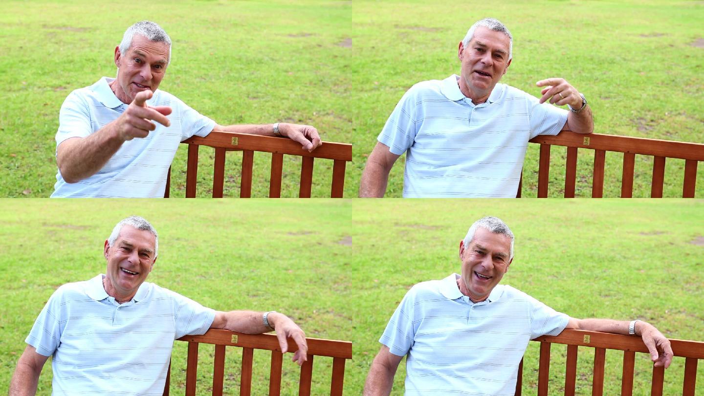 一个阳光明媚的日子里，一个退休男子坐在公园的长椅上对着摄像机说话