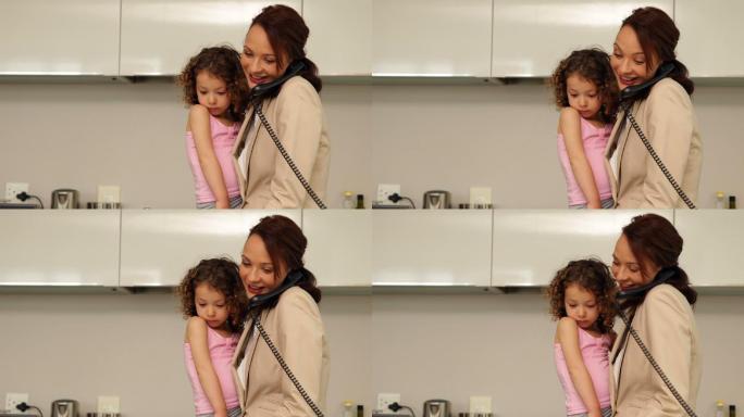 微笑的母亲抱着她的小女儿在家里的厨房打电话