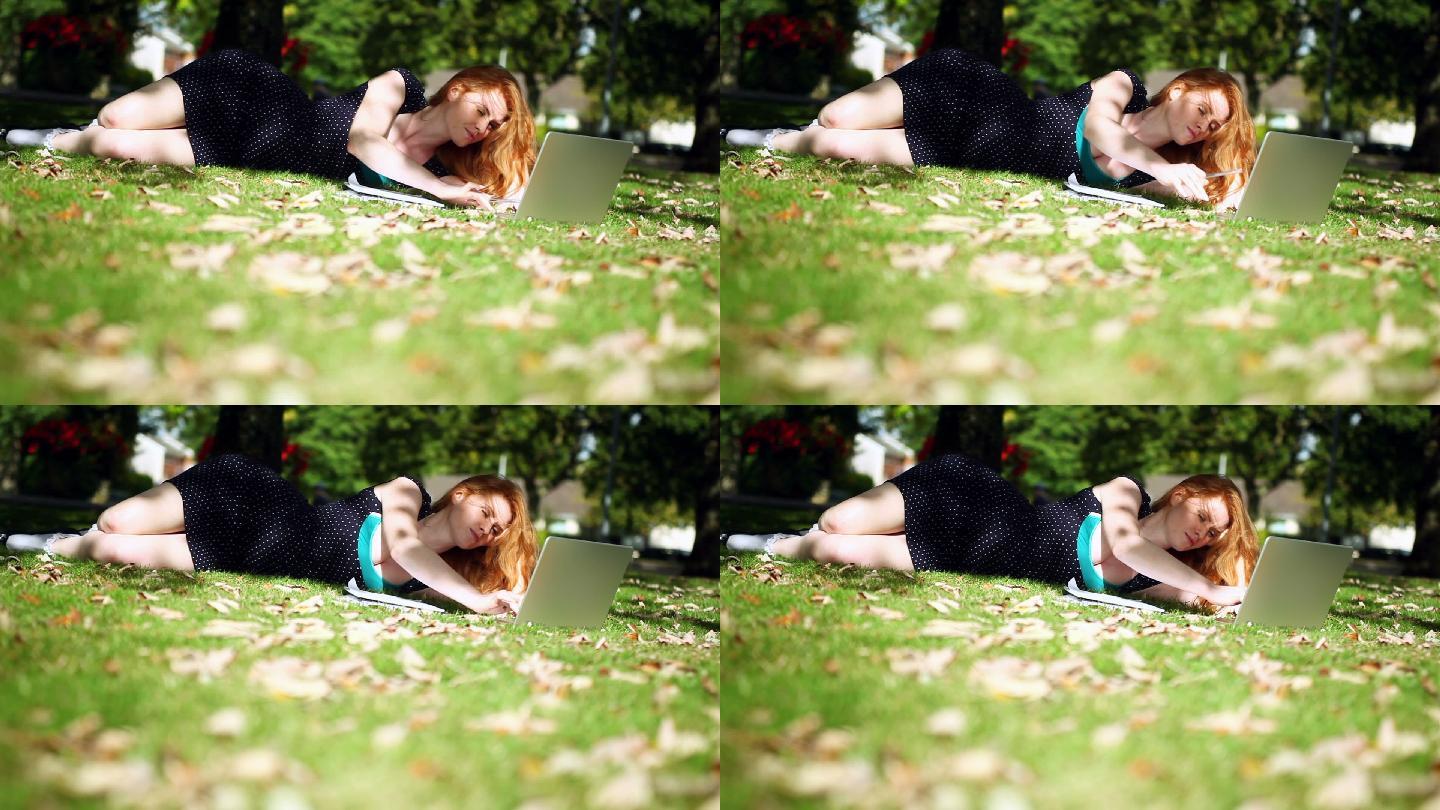 可爱的红发女孩躺在绿色的草坪上用她的笔记本做作业