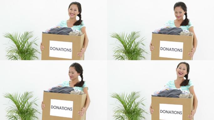漂亮的黑发女子拿着一个装满衣服的捐赠箱，对着镜头微笑