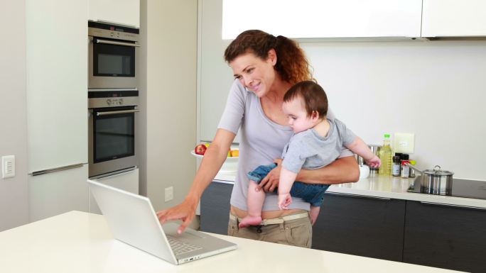 母亲抱着她快乐的宝宝，在厨房里用笔记本电脑打电话