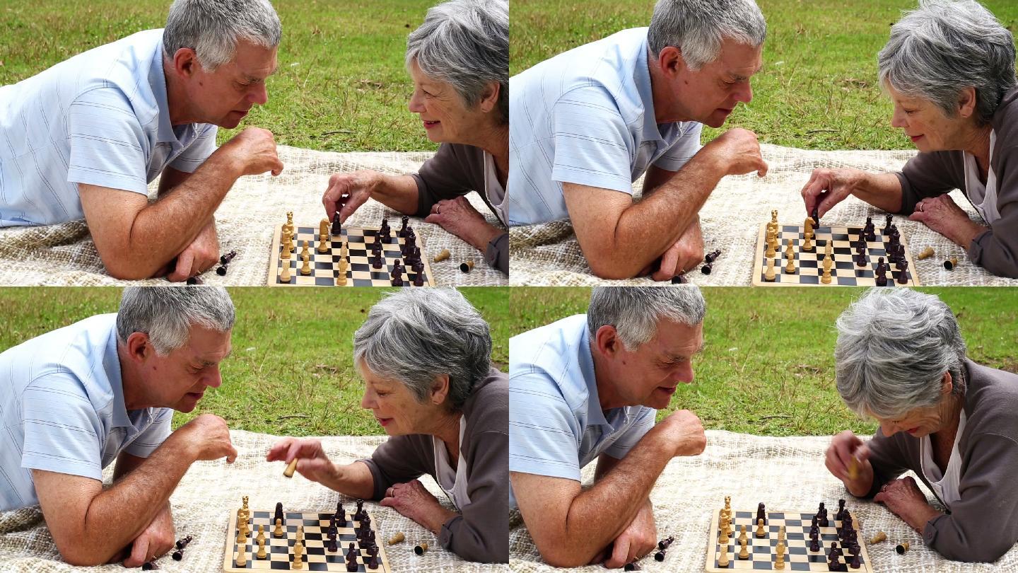 在一个阳光明媚的日子里，一对老年夫妇躺在公园里的毯子上下棋