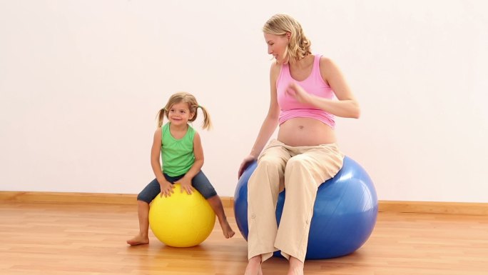 金发孕妇坐在健身球和她的小女儿在健身室