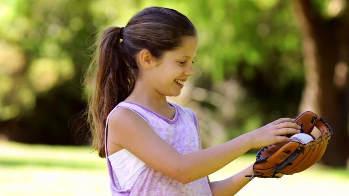 运动小女孩对着镜头微笑，在一个阳光明媚的日子里，在公园里玩棒球