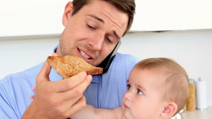一个商人抱着他的孩子，一边在家里的厨房里吃吐司一边打电话