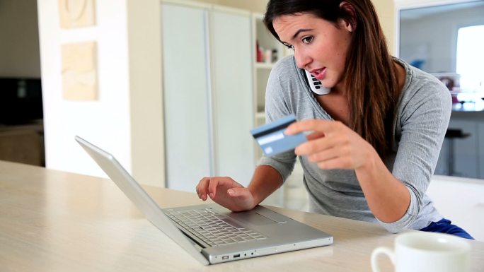 漂亮的黑发女人使用笔记本电脑在网上购物，同时在家里的厨房打电话