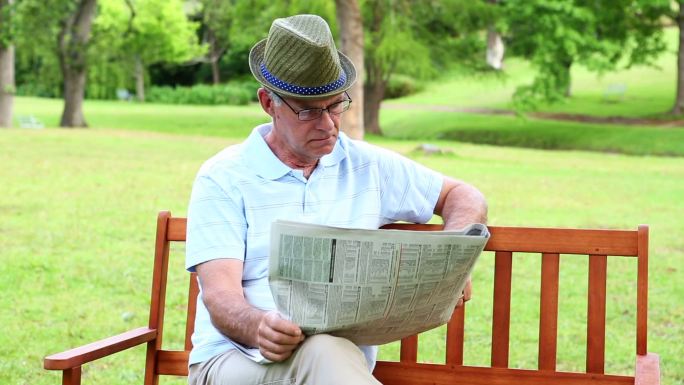 一个阳光明媚的日子，一个退休男子坐在公园的长椅上看报纸