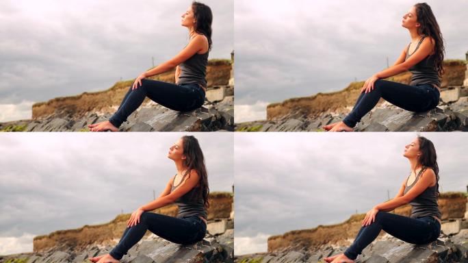 一个美丽的黑发女人坐在阴天的海滩边的岩石上