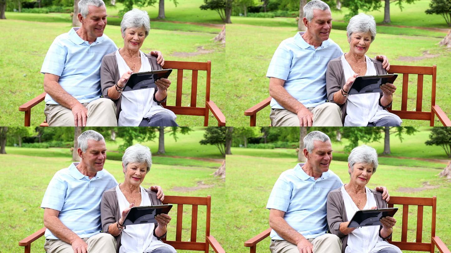 一对退休夫妇在一个阳光明媚的日子里坐在公园的长椅上使用平板电脑