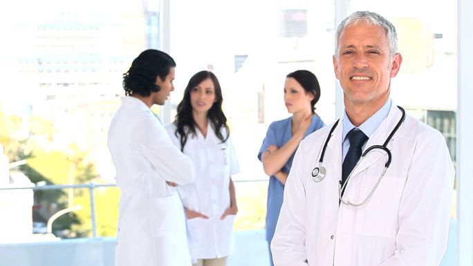 微笑着成熟的医生站在他的团队前面，在一个明亮的房间里