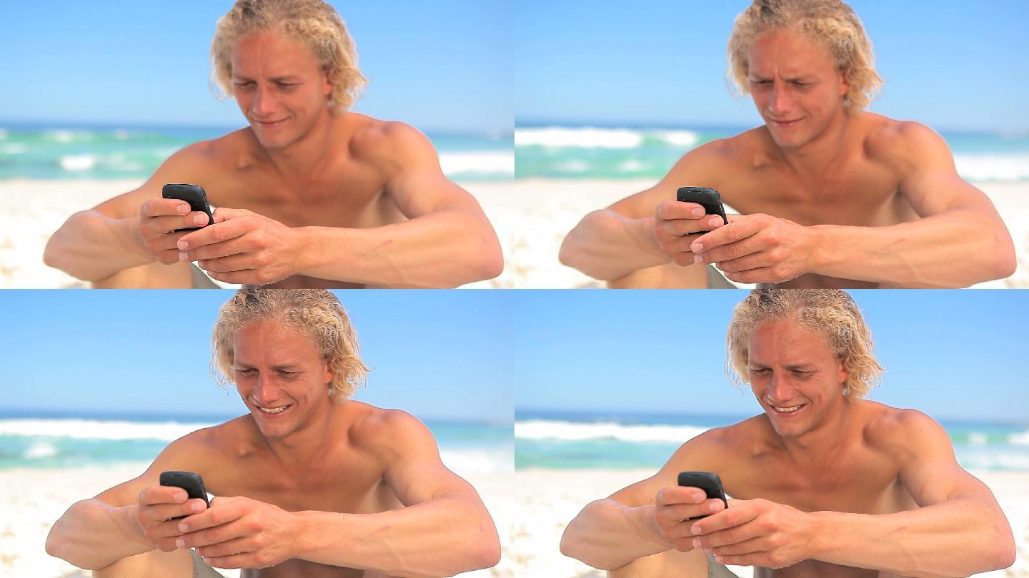 微笑的金发男人坐在沙滩上发短信