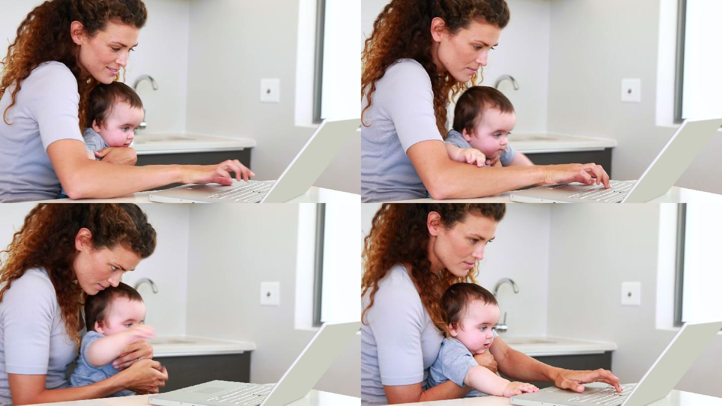 母亲和婴儿坐在膝盖上，在厨房里使用笔记本电脑