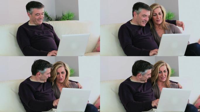 在家里的客厅里，一个男人在沙发上用着他的笔记本电脑，妻子坐在他旁边