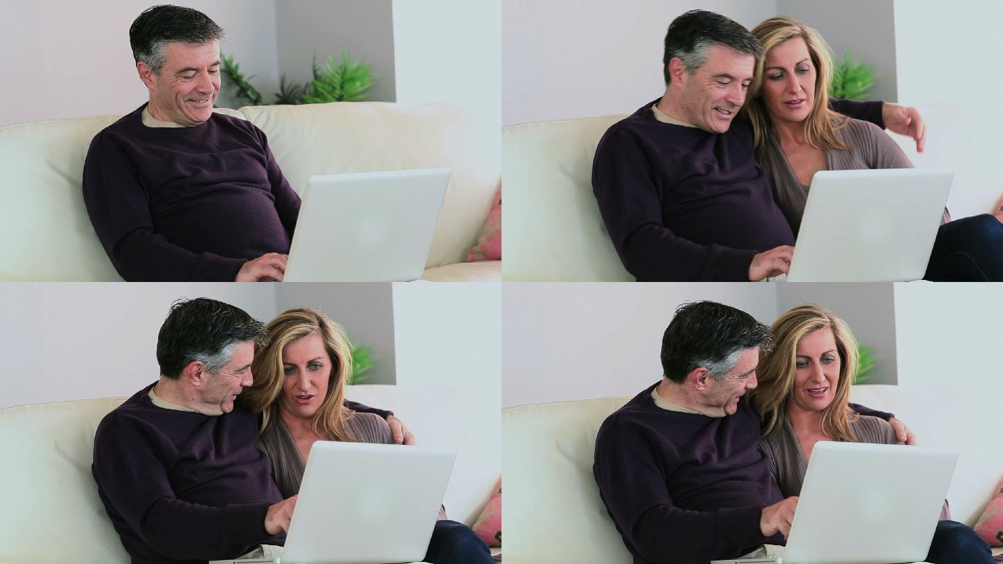 在家里的客厅里，一个男人在沙发上用着他的笔记本电脑，妻子坐在他旁边