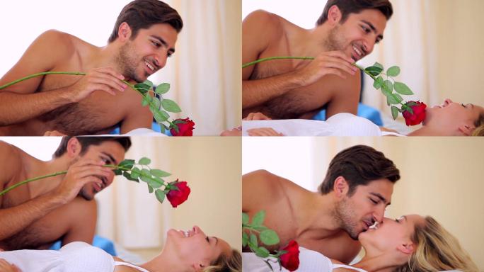 一个男人在卧室的床上用一朵红玫瑰给他漂亮的女朋友搔痒