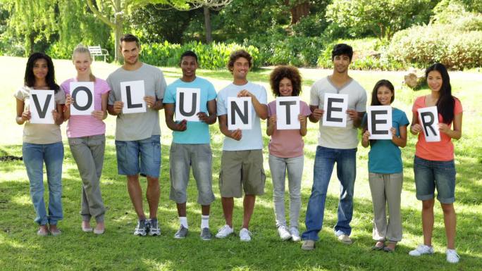 在一个阳光明媚的日子里，一群年轻的朋友在照相机前微笑着拿着字母拼写志愿者
