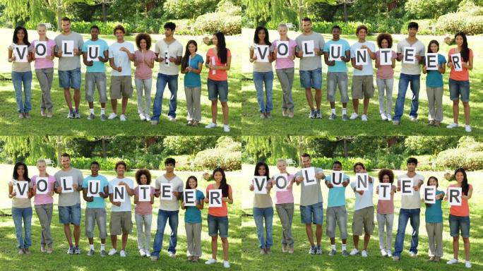在一个阳光明媚的日子里，一群年轻的朋友在照相机前微笑着拿着字母拼写志愿者