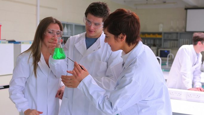 科学家们在实验室里分组研究绿色液体的烧杯，他们的老师过来给他们讲解