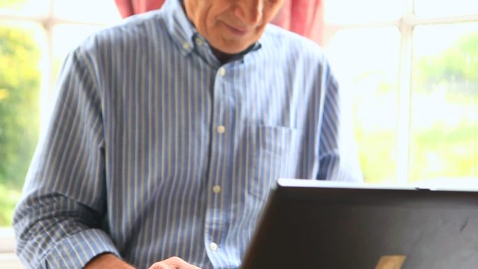 专注的成熟男子坐在窗边使用他的电脑在他的客厅