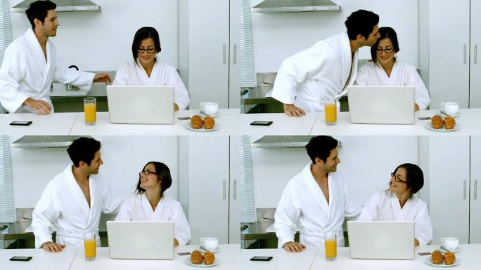 一个男人亲吻他的女朋友，她正在用她的笔记本电脑在厨房吃早餐