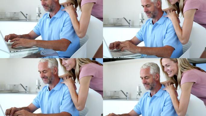 一个男人在笔记本电脑上打字，而他的妻子微笑着拥抱着他