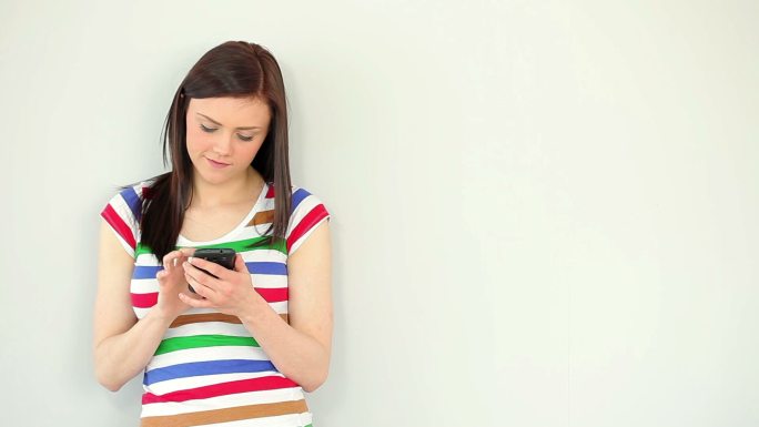 一名年轻女子面带微笑，靠在白墙上用手机发短信