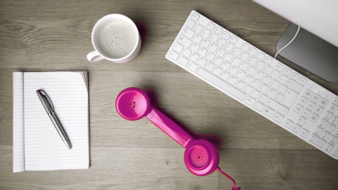 粉红色的电话听筒掉到桌子上，旁边是电脑、咖啡和记事本