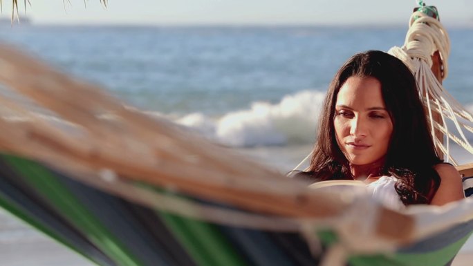 可爱的黑发女人在沙滩上的吊床上看书