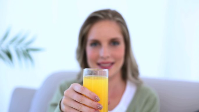 一个女人坐在沙发上，向镜头展示她的一杯橙汁
