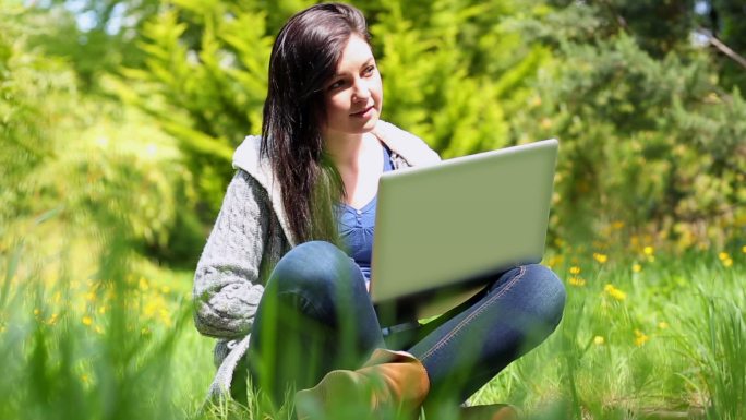 快乐的年轻女子坐在草地上使用笔记本电脑在阳光明媚的日子