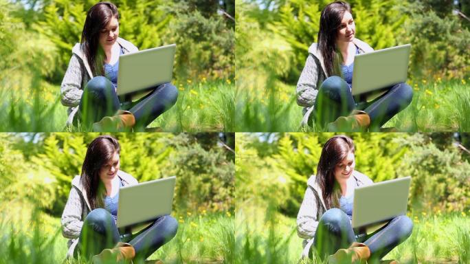 快乐的年轻女子坐在草地上使用笔记本电脑在阳光明媚的日子