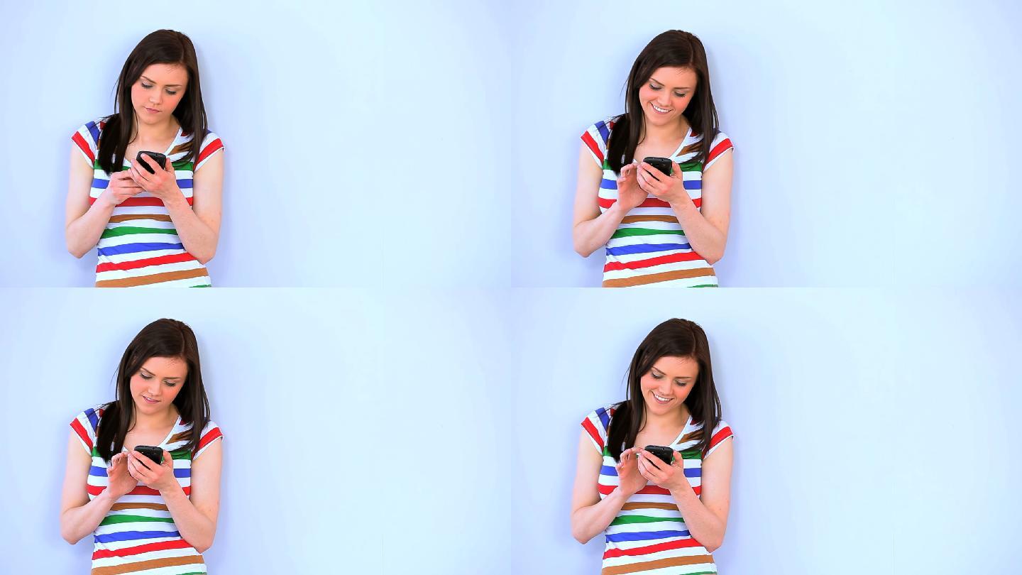 一名黑发女子靠在白墙上用智能手机发短信