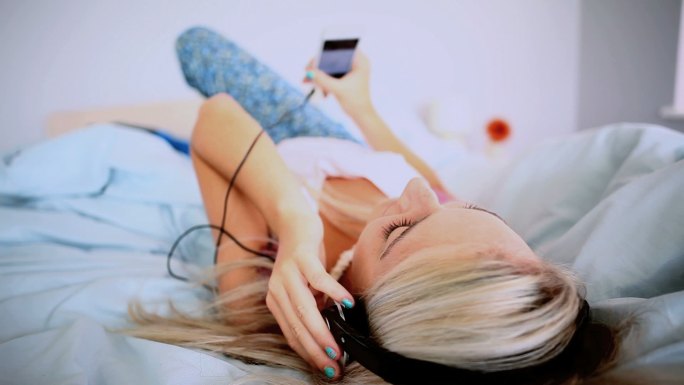 迷人的金发女郎躺在床上，在明亮的卧室里听音乐