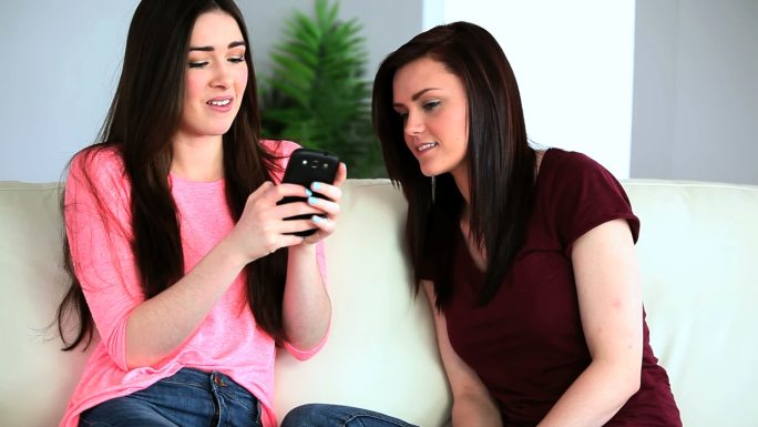 一位黑发女子向坐在沙发上的朋友展示她的智能手机