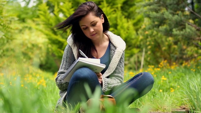 一个有风的日子，微笑的年轻女子坐在草地上写在记事本上