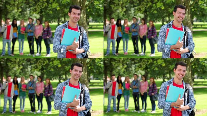 在大学校园的草地上，一个学生对着镜头微笑，他的朋友站在他身后