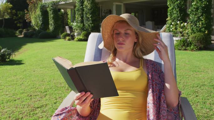 一个白人妇女坐在花园的躺椅上一边看书一边晒日光浴
