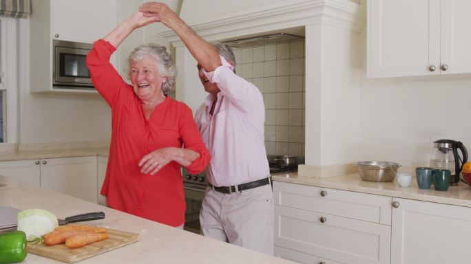 快乐的白种人老夫妇有乐趣在厨房跳舞，同时准备晚餐
