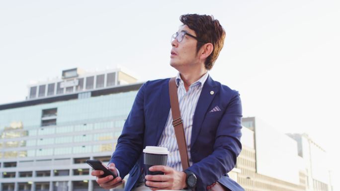 一名亚洲男子坐在企业园区的屋顶上喝着咖啡，用着智能手机
