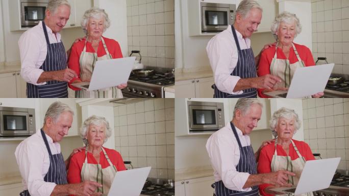 快乐的白种人老夫妇在厨房穿着围裙使用笔记本电脑准备饭前