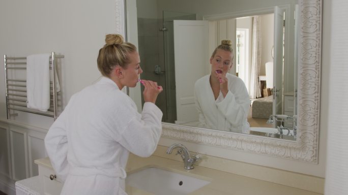 穿着浴衣的白人妇女在浴室里一边看着镜子一边刷牙