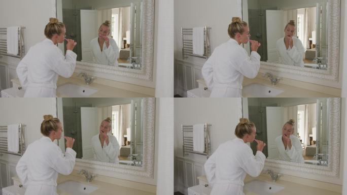 穿着浴衣的白人妇女在浴室里一边看着镜子一边刷牙