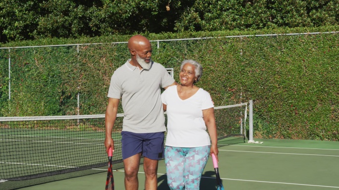 一对非洲裔美国老年夫妇拿着球拍在网球场上行走